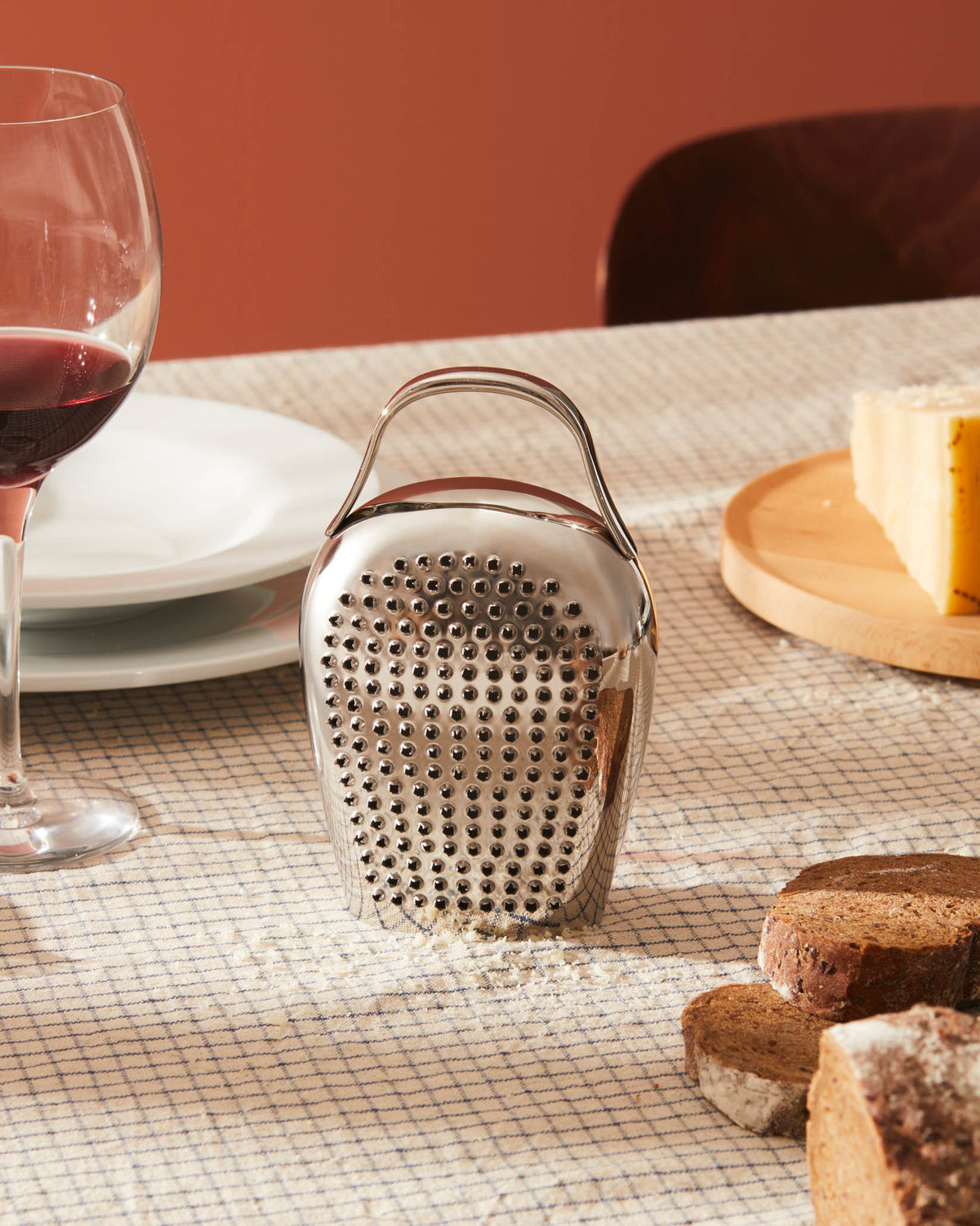 Silver bell-shaped grater by Alessi with a handle, surrounded by grated cheese on a checkered tablecloth.