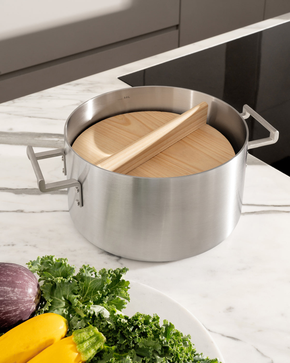 Natural wood Convivio lid by Alessi, split design, partially covering a stainless steel pot on a marble counter with fresh vegetables.
