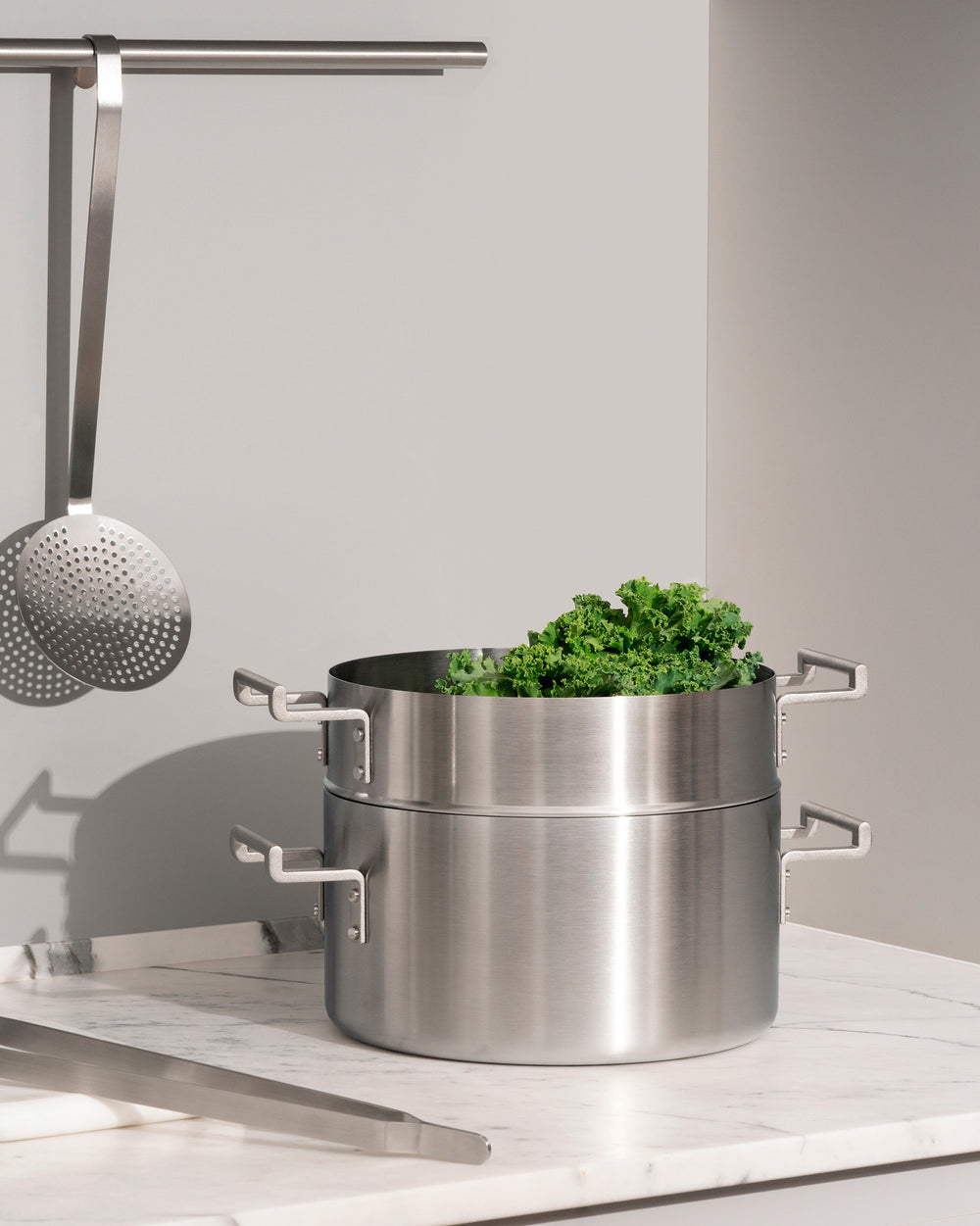 Two-tier brushed stainless steel steamer basket set by Alessi, filled with fresh green kale, on a white marble countertop.