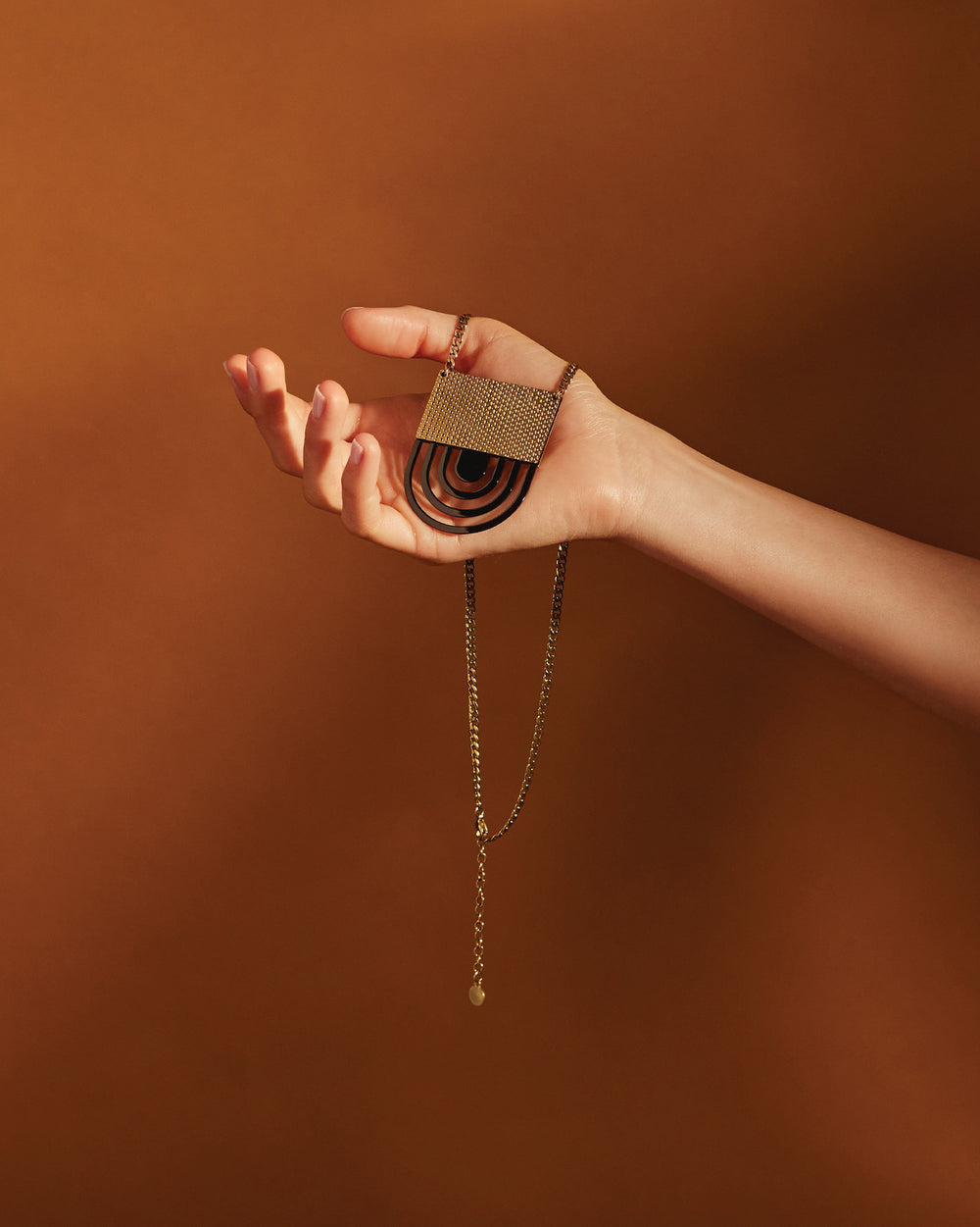 Gold-toned necklace by Alessi featuring a textured rectangular top and black concentric arch pendant, held in a hand.