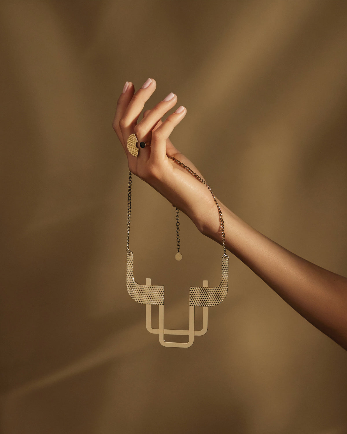 Textured gold-toned geometric necklace by Alessi, held by a hand wearing a patterned ring against a brown background.