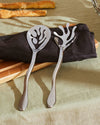 Shiny silver Mediterraneo salad servers by Alessi with organic, coral-like cutouts, resting on a dark napkin and wooden board.
