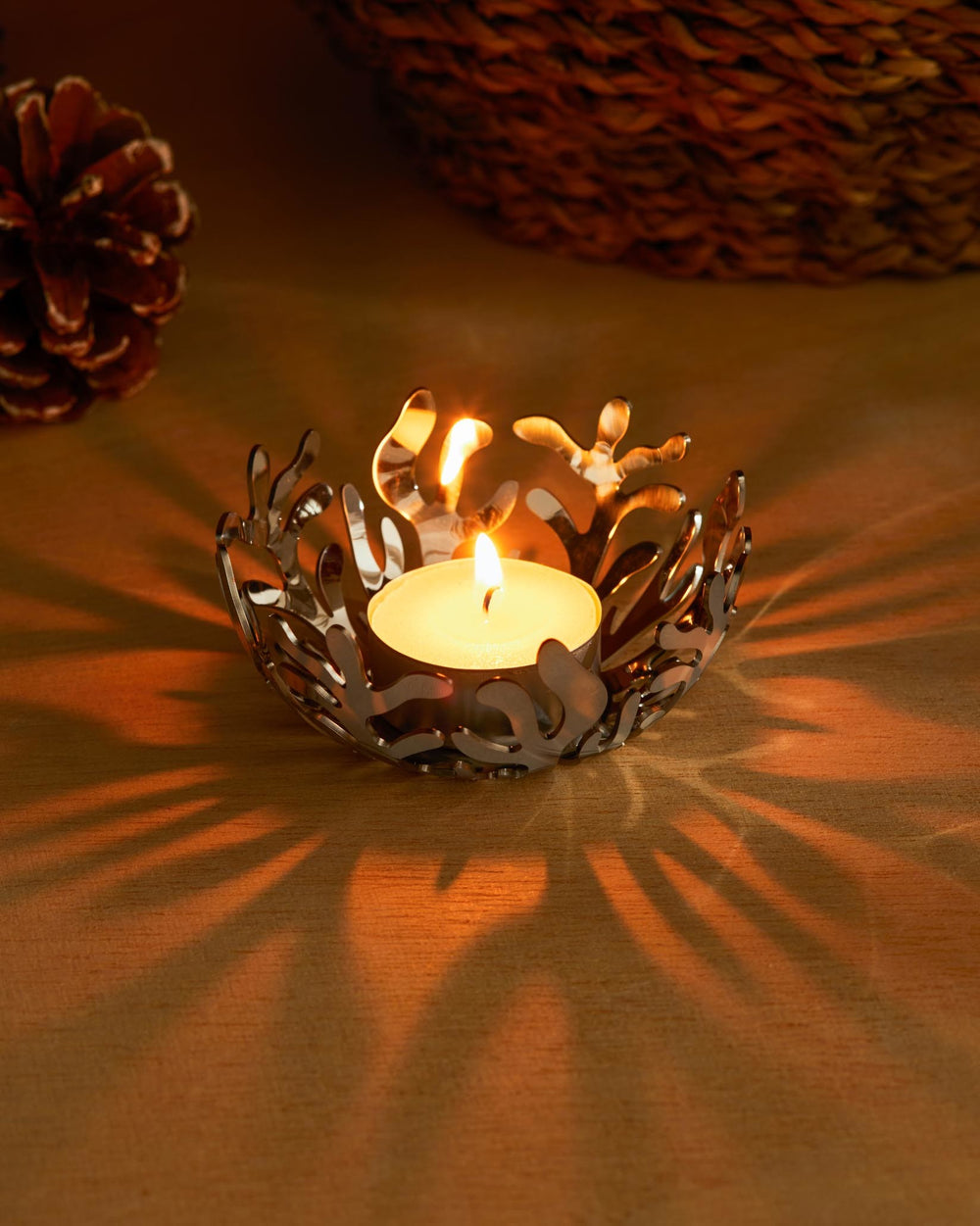 A silver metal tealight holder by Alessi with organic coral-like cutouts, casting elaborate shadows on a wooden surface.