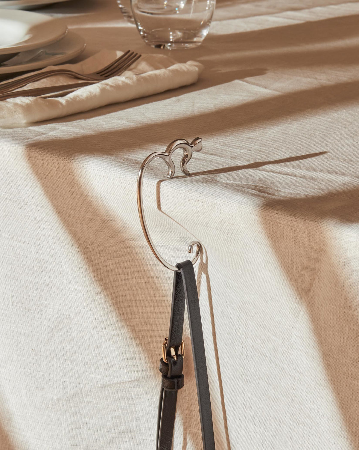 Silver-toned metal handbag hook by Alessi, cat-shaped, holding a black leather bag strap on a linen tablecloth.