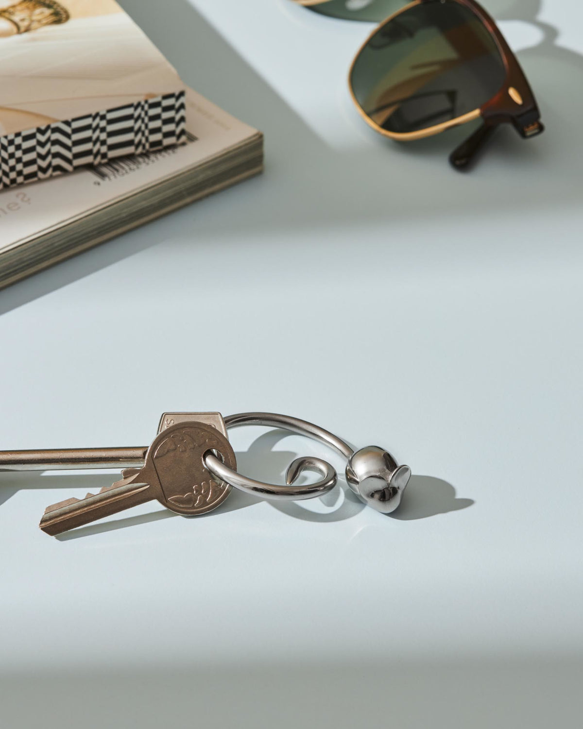 Silver-toned mouse-shaped keychain by Alessi, its reflective metal surface lying on a light blue table.