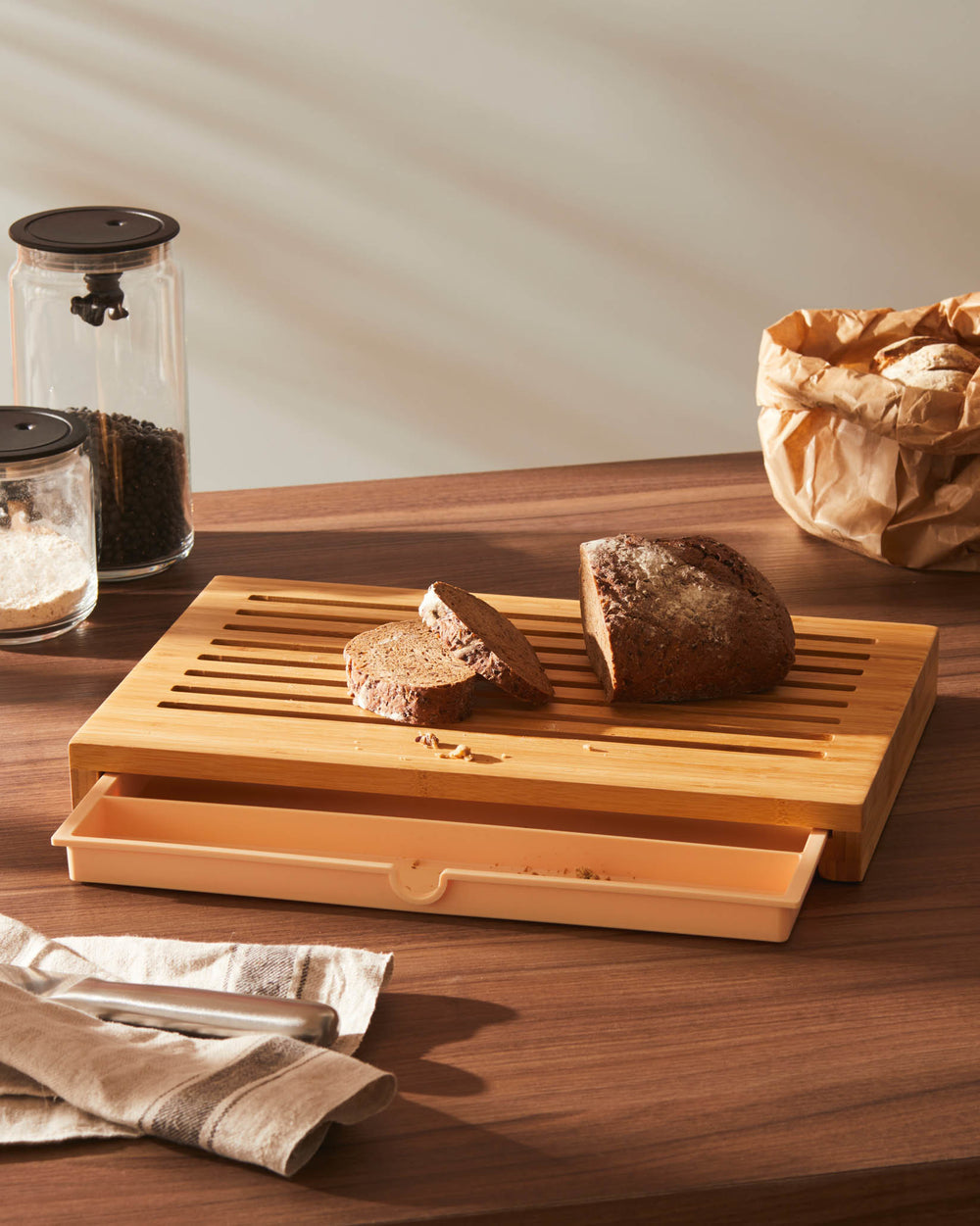 Light wood chopping board with crumb tray by Alessi, holding dark bread slices on a wooden countertop.