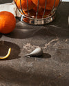 Sleek silver-grey citrus peeler by Alessi, positioned on a speckled dark surface with an orange and peel nearby.