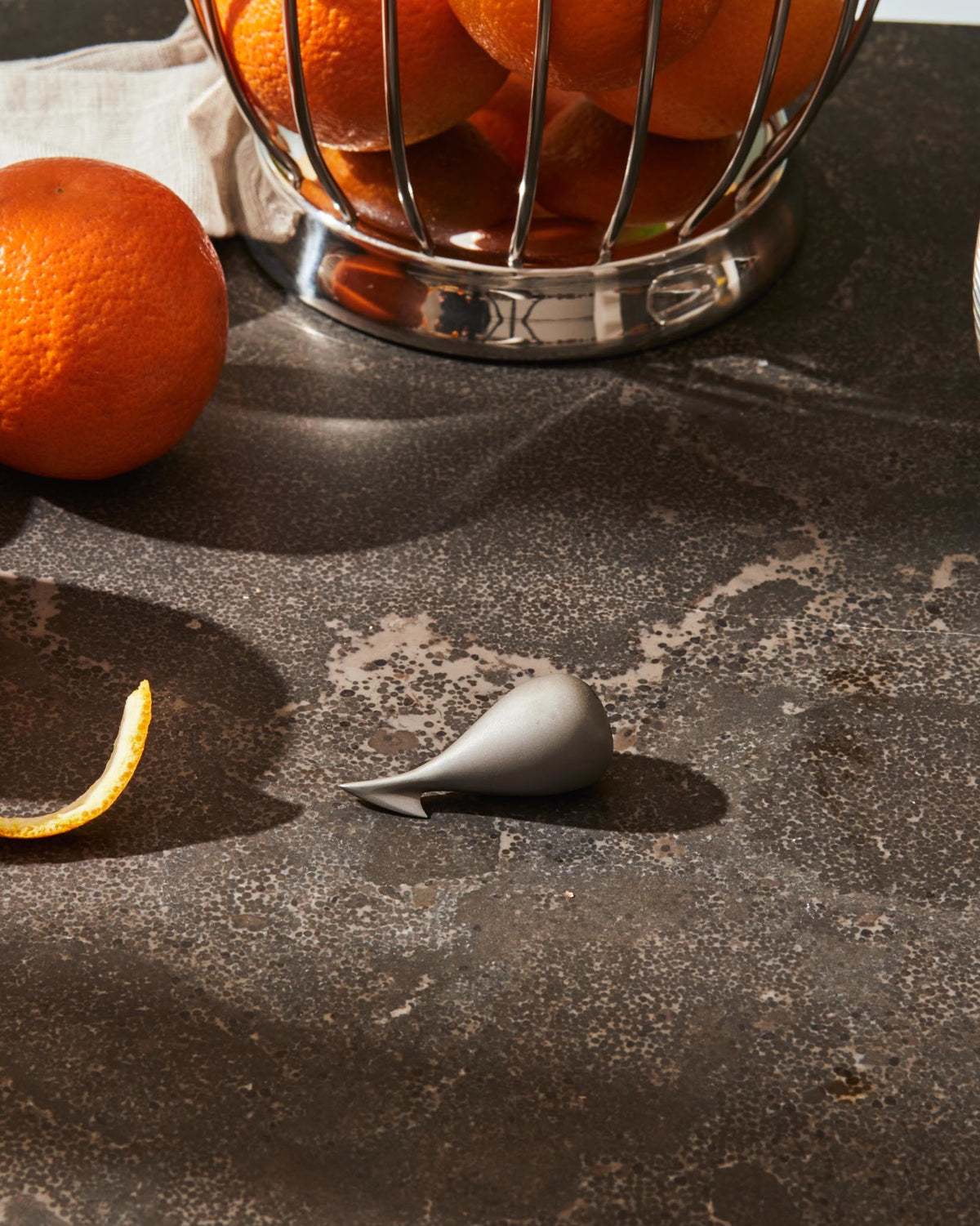 Sleek silver-grey citrus peeler by Alessi, positioned on a speckled dark surface with an orange and peel nearby.