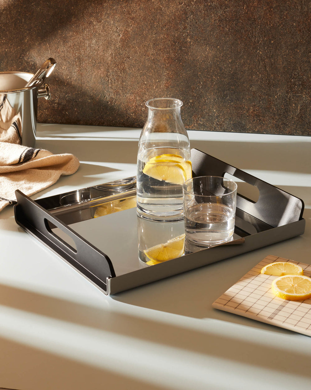 A dark grey and polished stainless steel serving tray by Alessi, on a light blue countertop, holding a carafe and glass of lemon water.