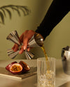 The tending box by Alessi, featuring a stainless steel jigger pouring golden liquid into a fluted glass with ice and passion fruit.