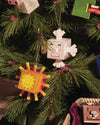 White cube-shaped Snowray ornament by Alessi, with a smiling face, hanging on a Christmas tree next to an orange sun ornament.