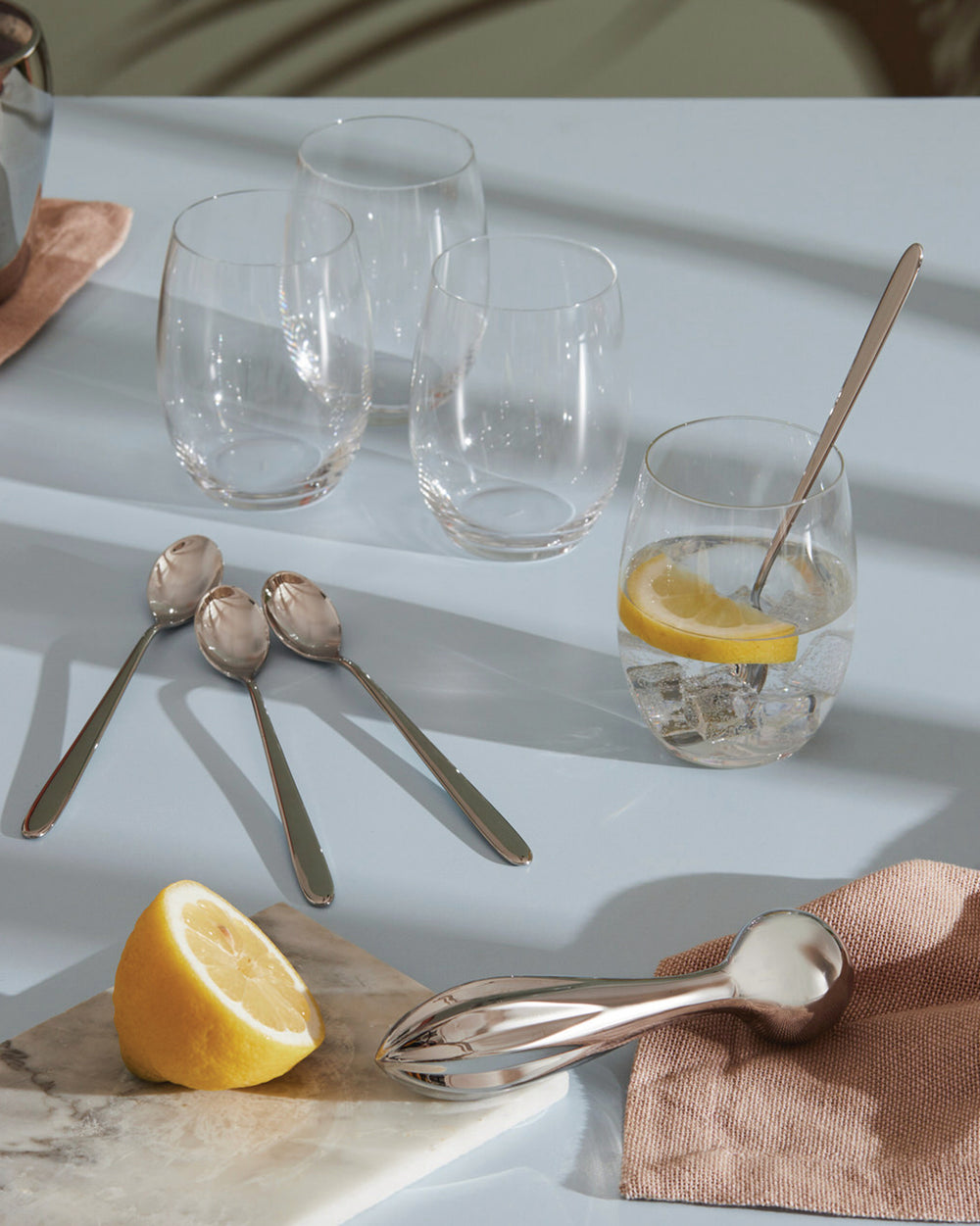 Silver lemon squeezer by Alessi on a white marble board with a half lemon, spoons, and drinks.
