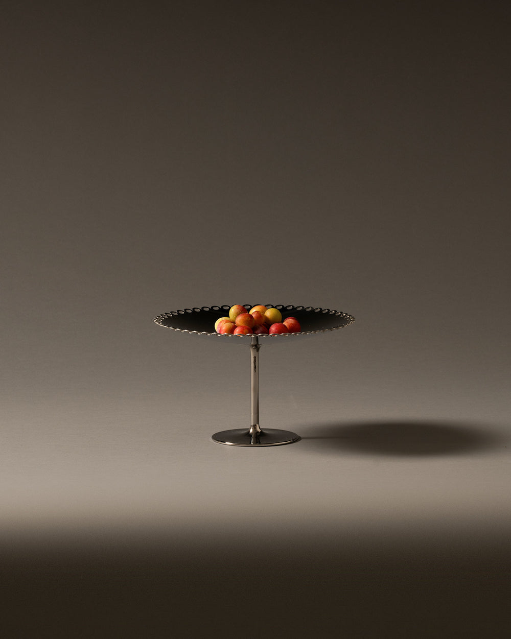 Silver and dark fruit stand with a scalloped edge by Alessi, holding colorful fruit.