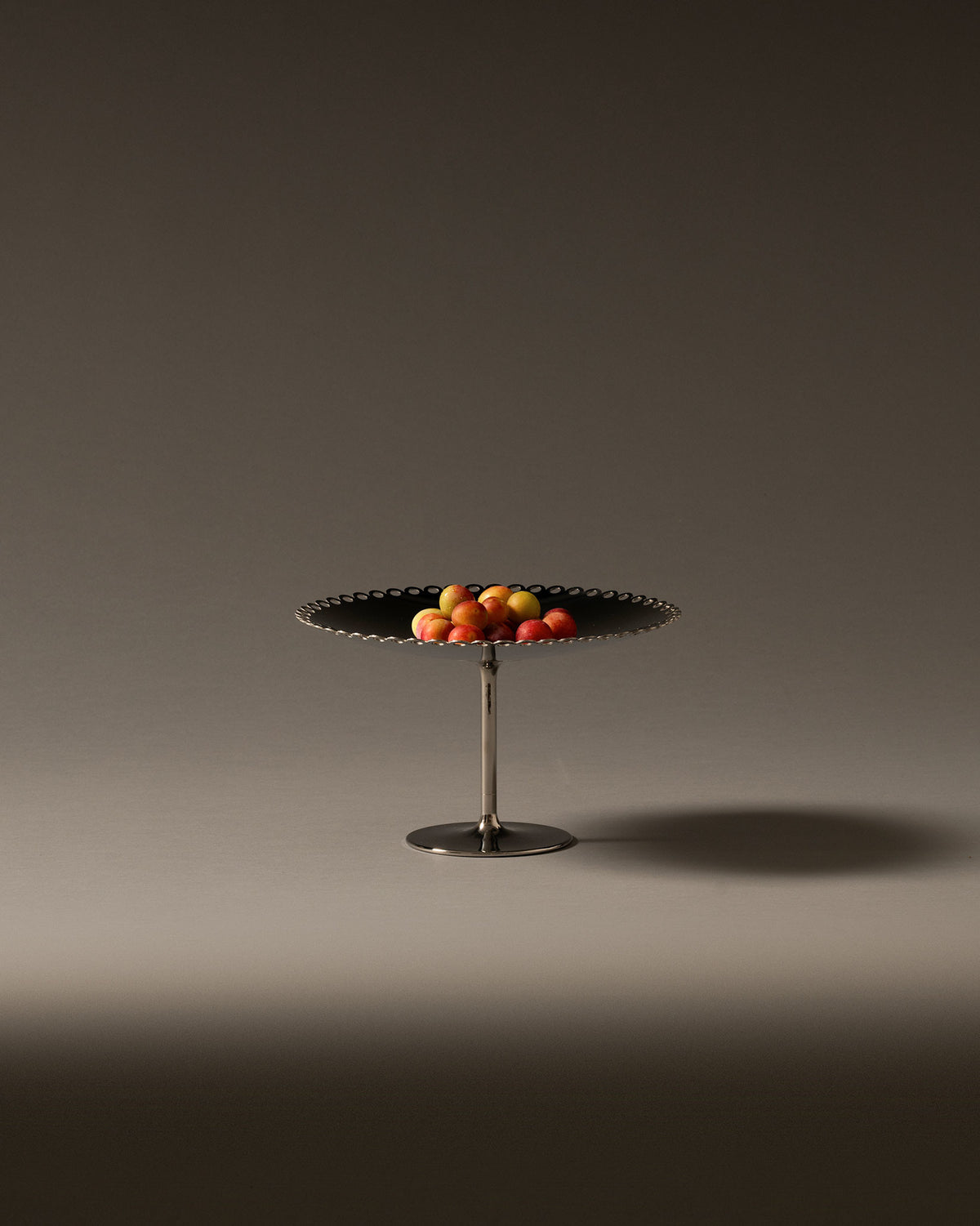 Silver and dark fruit stand with a scalloped edge by Alessi, holding colorful fruit.