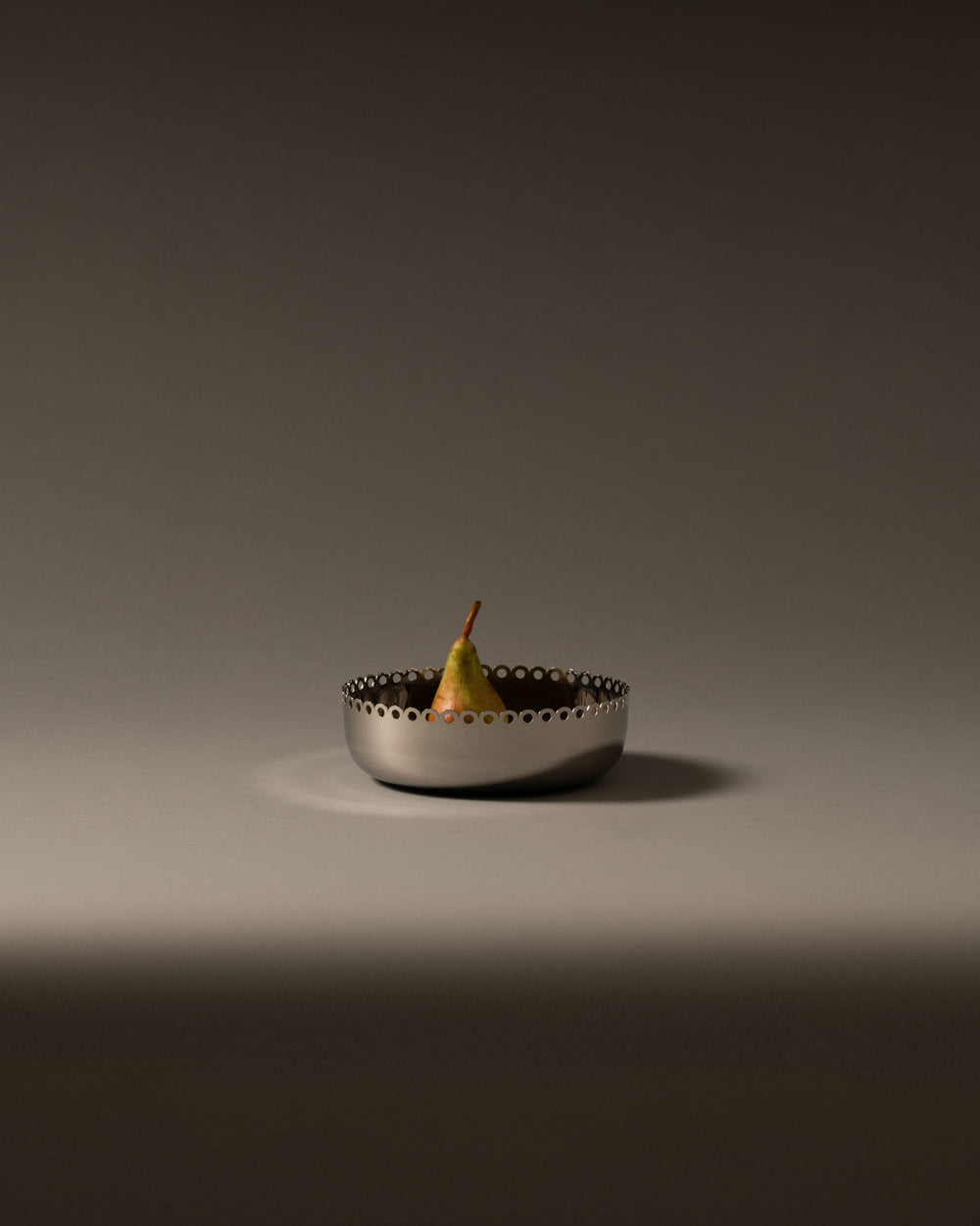 Silver fruit bowl by Alessi with a perforated scalloped edge, holding a single pear.