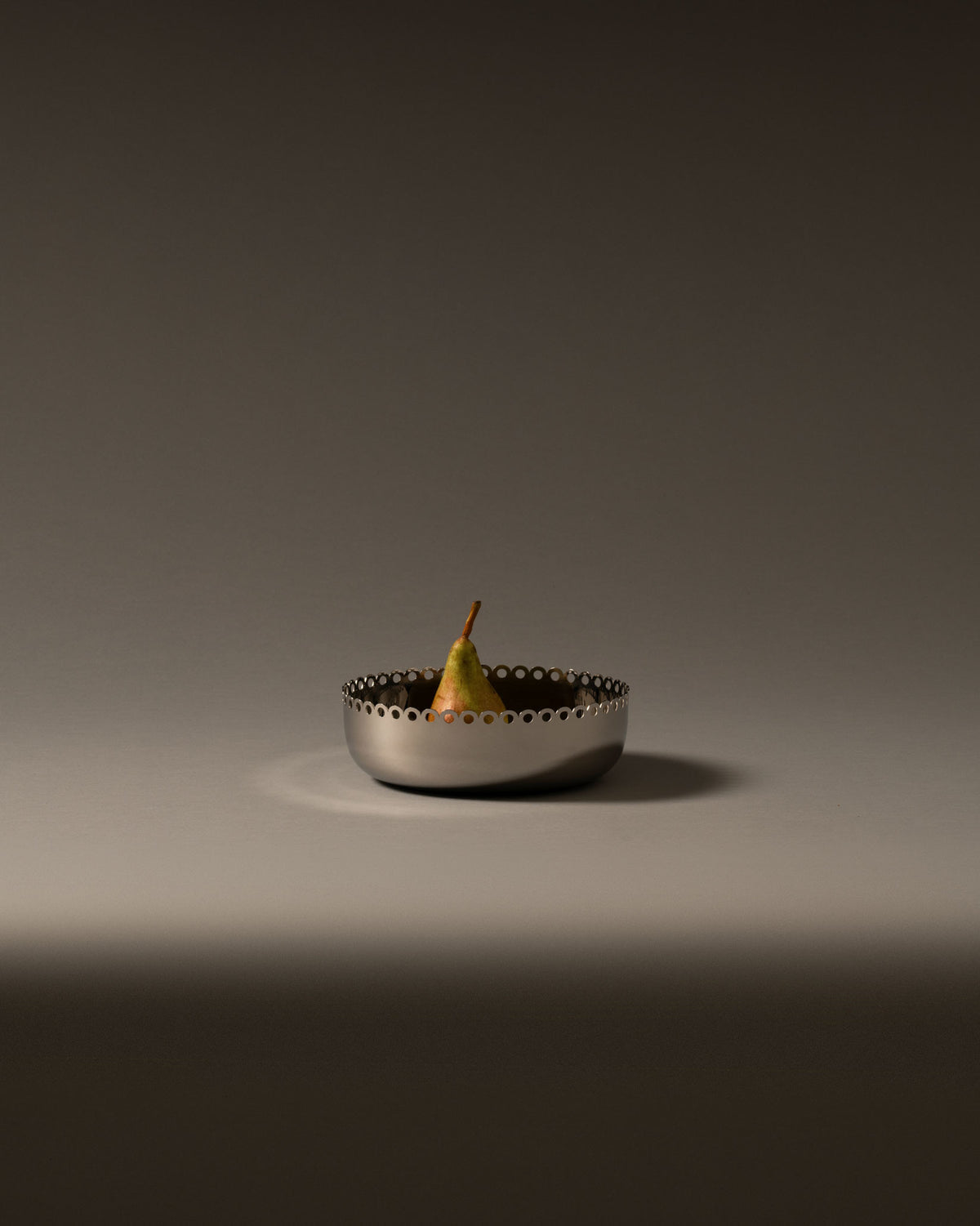 Silver fruit bowl by Alessi with a perforated scalloped edge, holding a single pear.