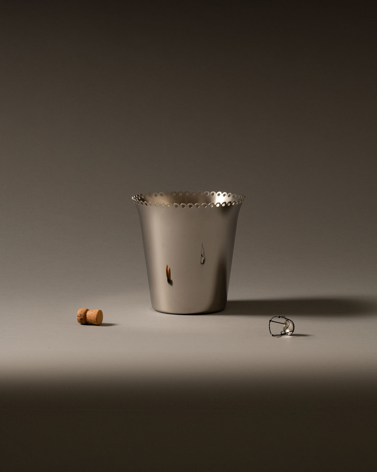 Silver wine cooler by Alessi with a perforated top edge, displayed with a cork and wire cage on a grey surface.