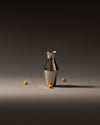 Shiny metallic pitcher by Alessi, standing on a dark grey surface with several small yellow spheres around it.