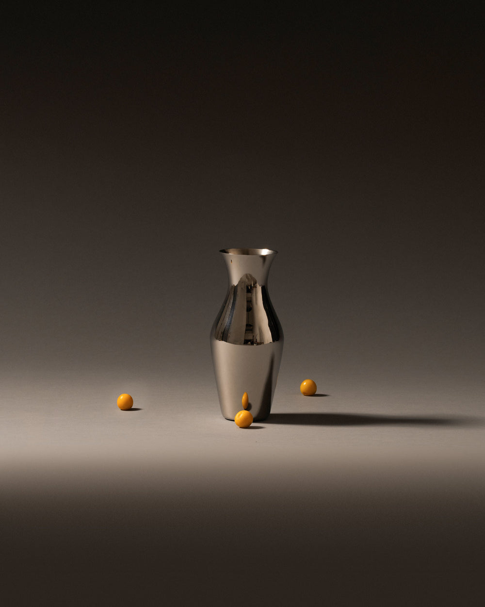 Shiny metallic pitcher by Alessi, standing on a dark grey surface with several small yellow spheres around it.