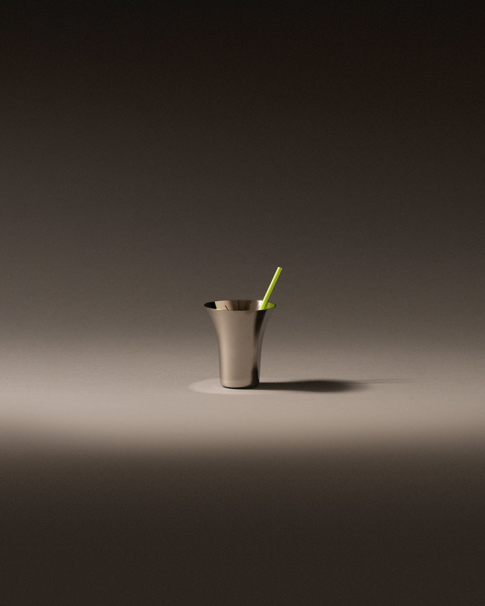 Brushed metal glass by Alessi with a flared rim and a green straw, on a grey surface.