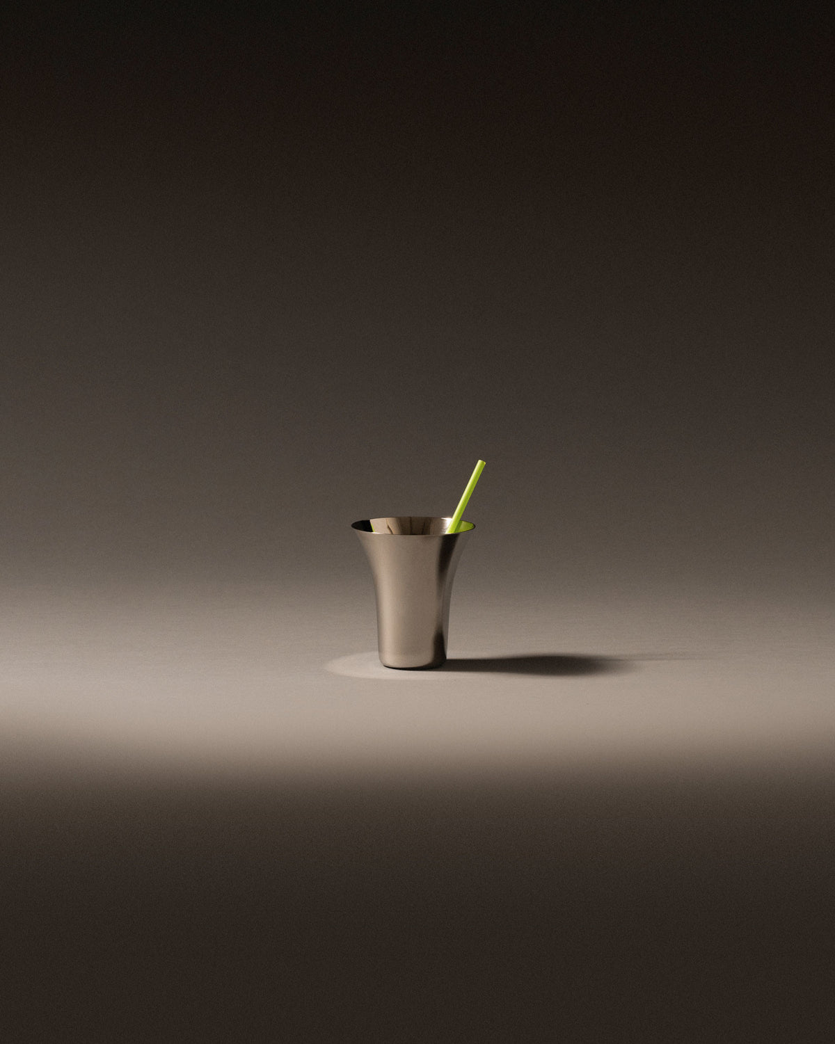 Brushed metal glass by Alessi with a flared rim and a green straw, on a grey surface.