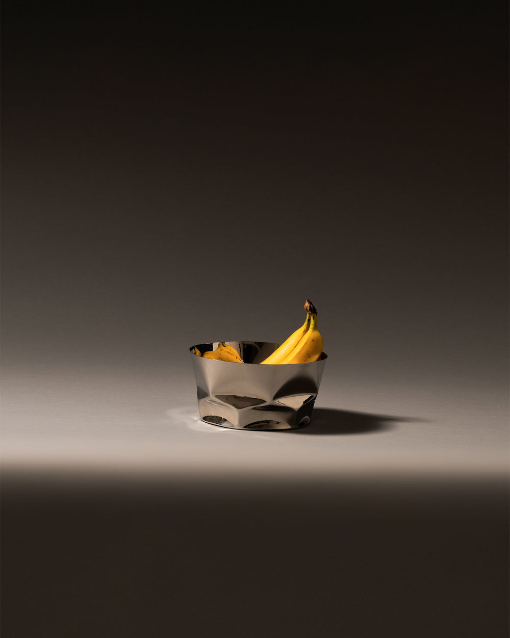 Silver stainless steel fruit bowl by Alessi with a crinkled design, holding two ripe bananas.