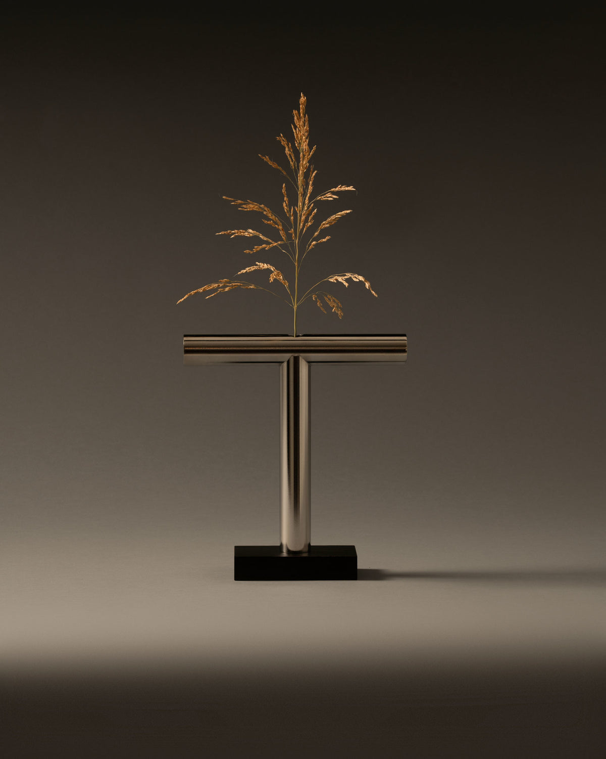 T-shaped metallic vase by Alessi on a black base, holding a dried golden plant.