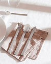Minimalist silver flatware set by Alessi, featuring a fork, spoon, and knife on a textured brown napkin.