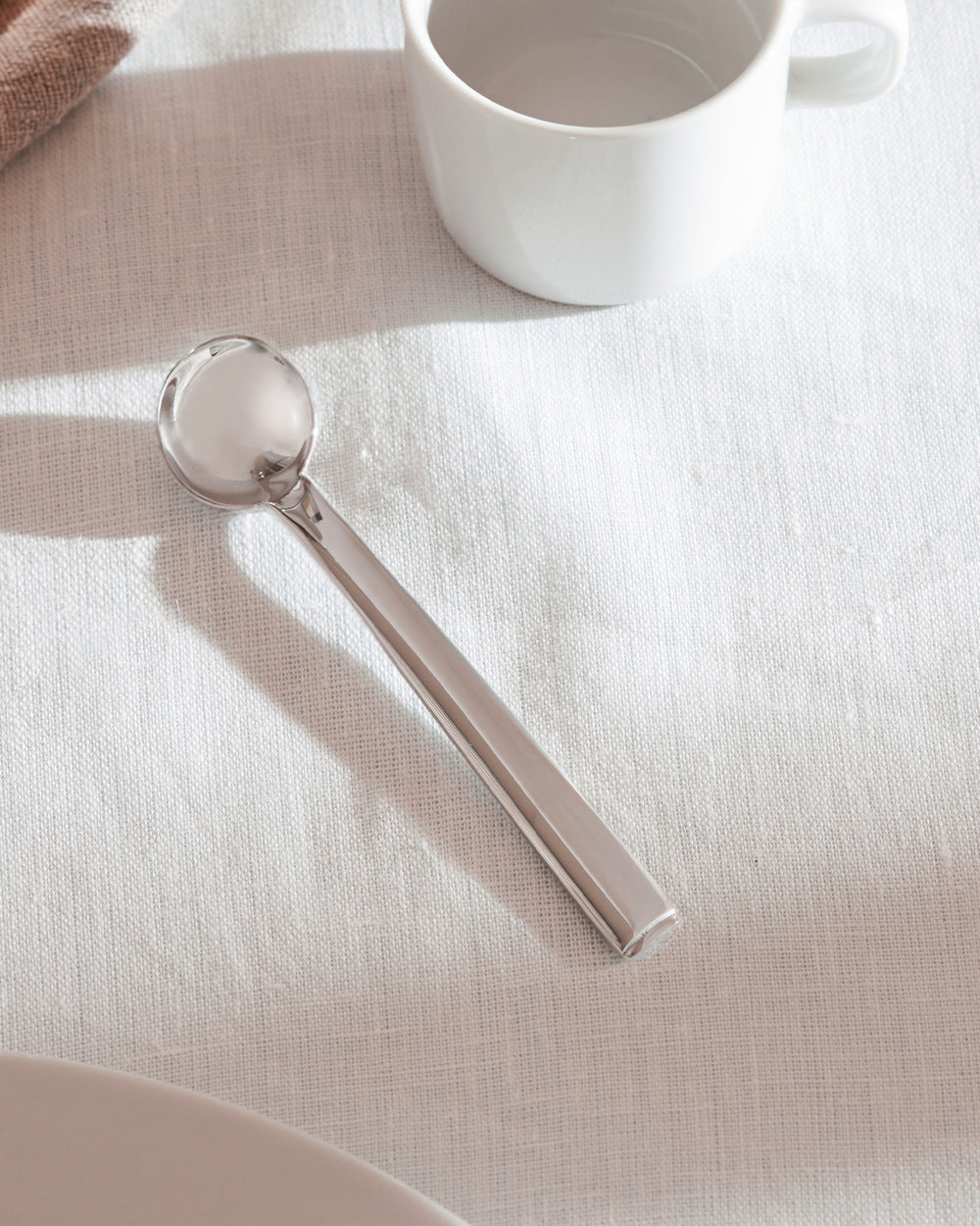 Silver round-bowled spoon by Alessi, polished metal, lying on a white linen tablecloth beside a white mug.