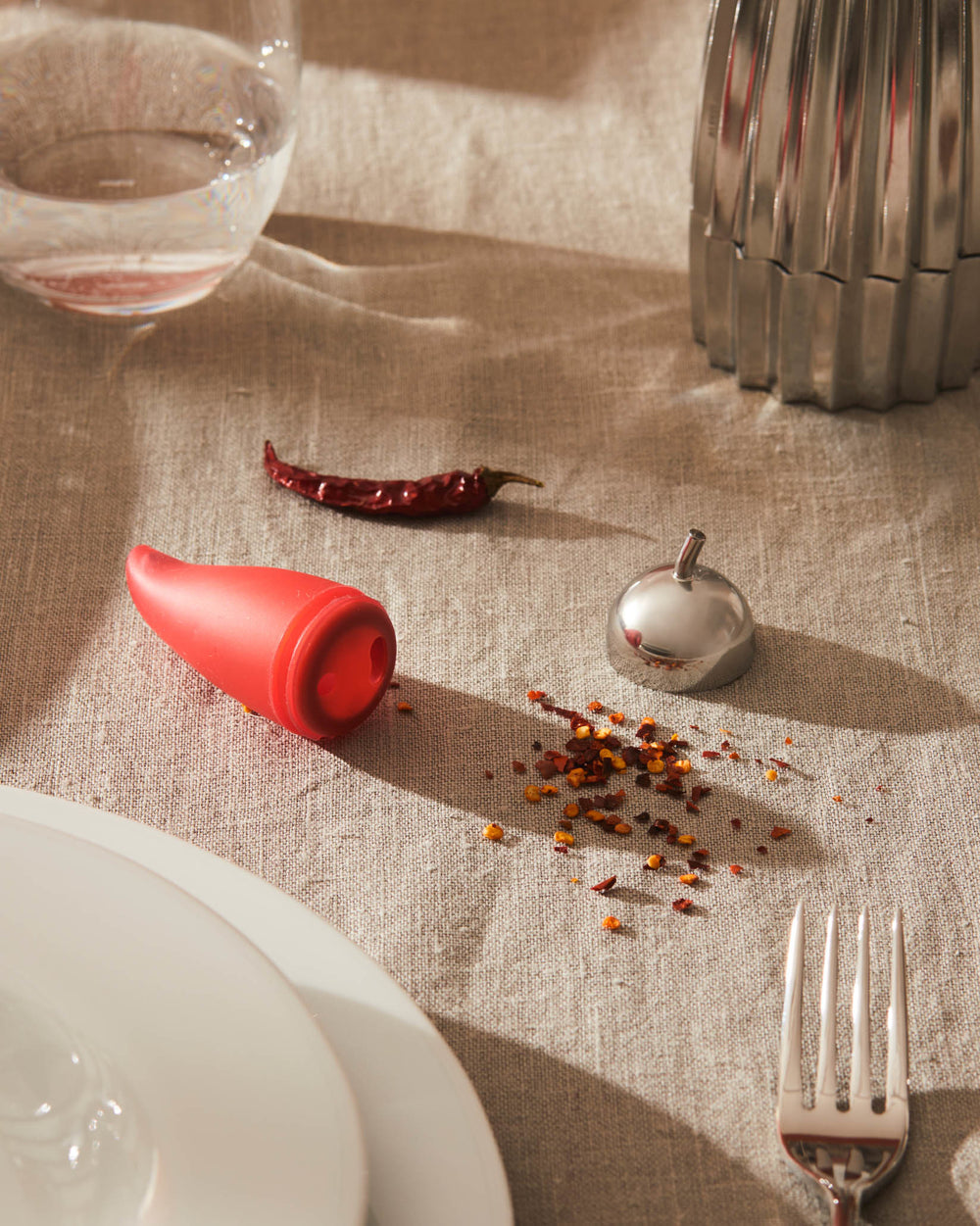 Red chili-shaped mill by Alessi with silver top, surrounded by chili flakes on a linen table.