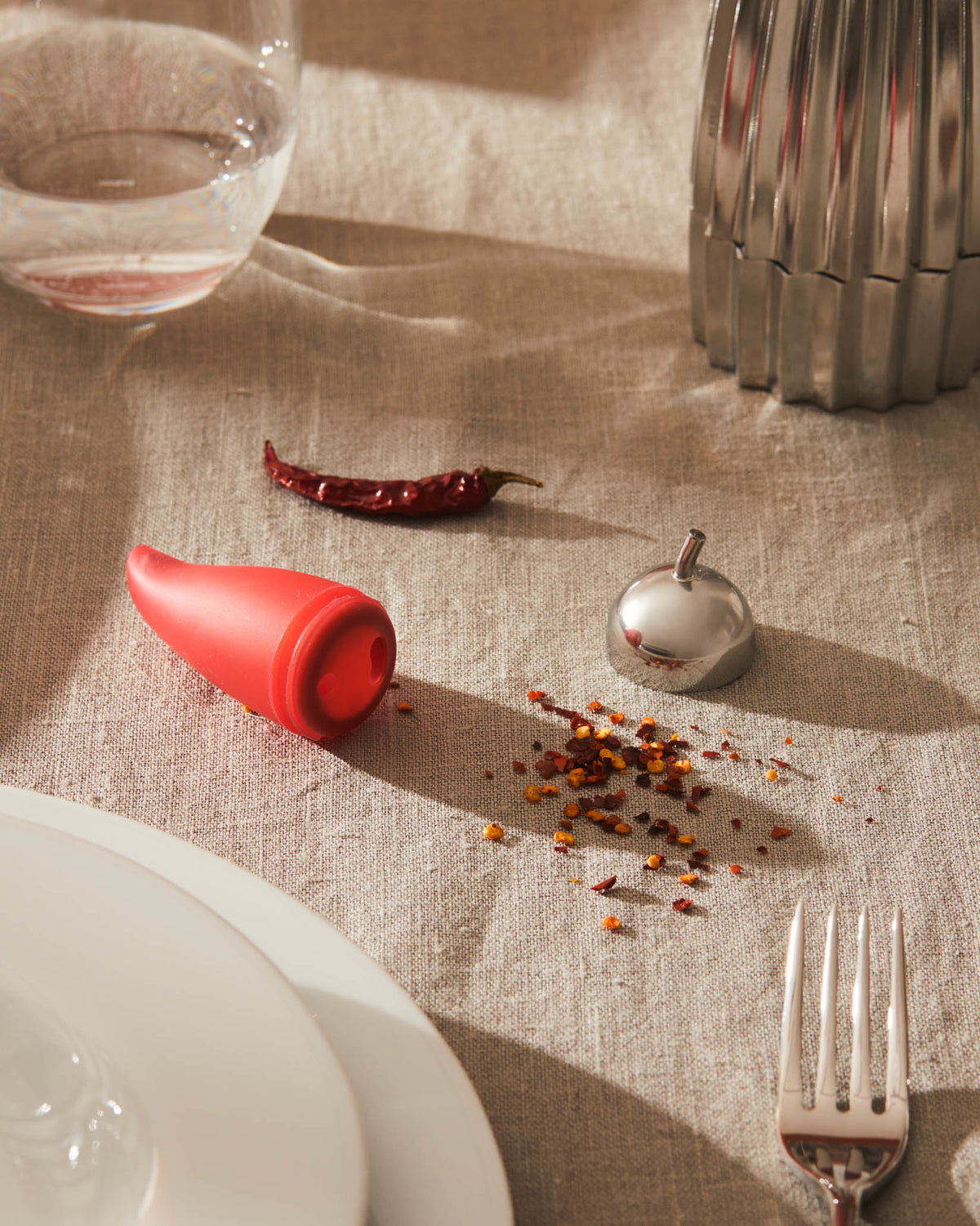 Red chili-shaped mill by Alessi with silver top, surrounded by chili flakes on a linen table.