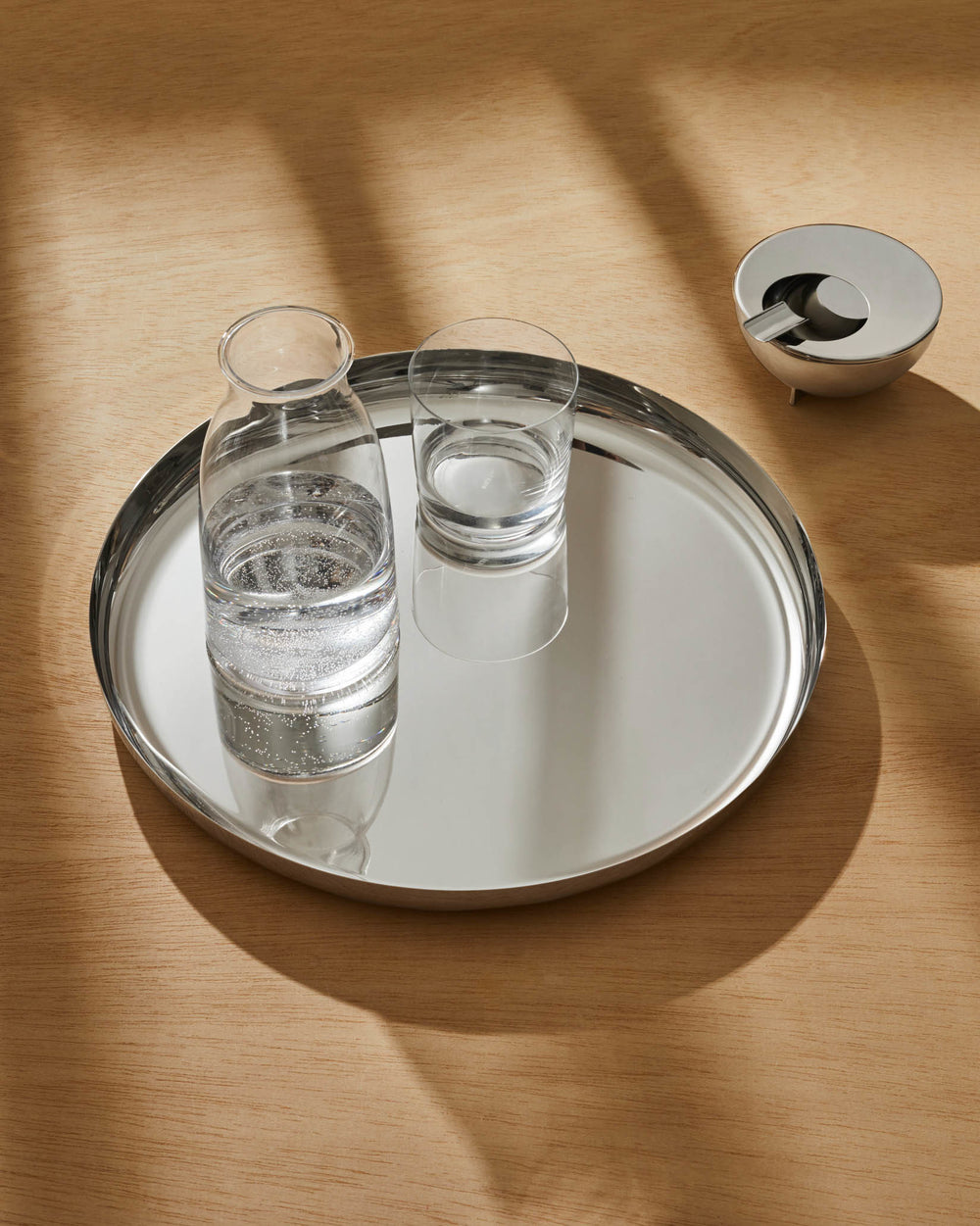 A round silver serving tray by Alessi holding a glass carafe and drinking glass, with a metal ashtray on a wooden table.
