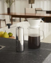 Chrome manual coffee grinder by Alessi with black handle, shown next to a glass pour-over coffee carafe and fruit.