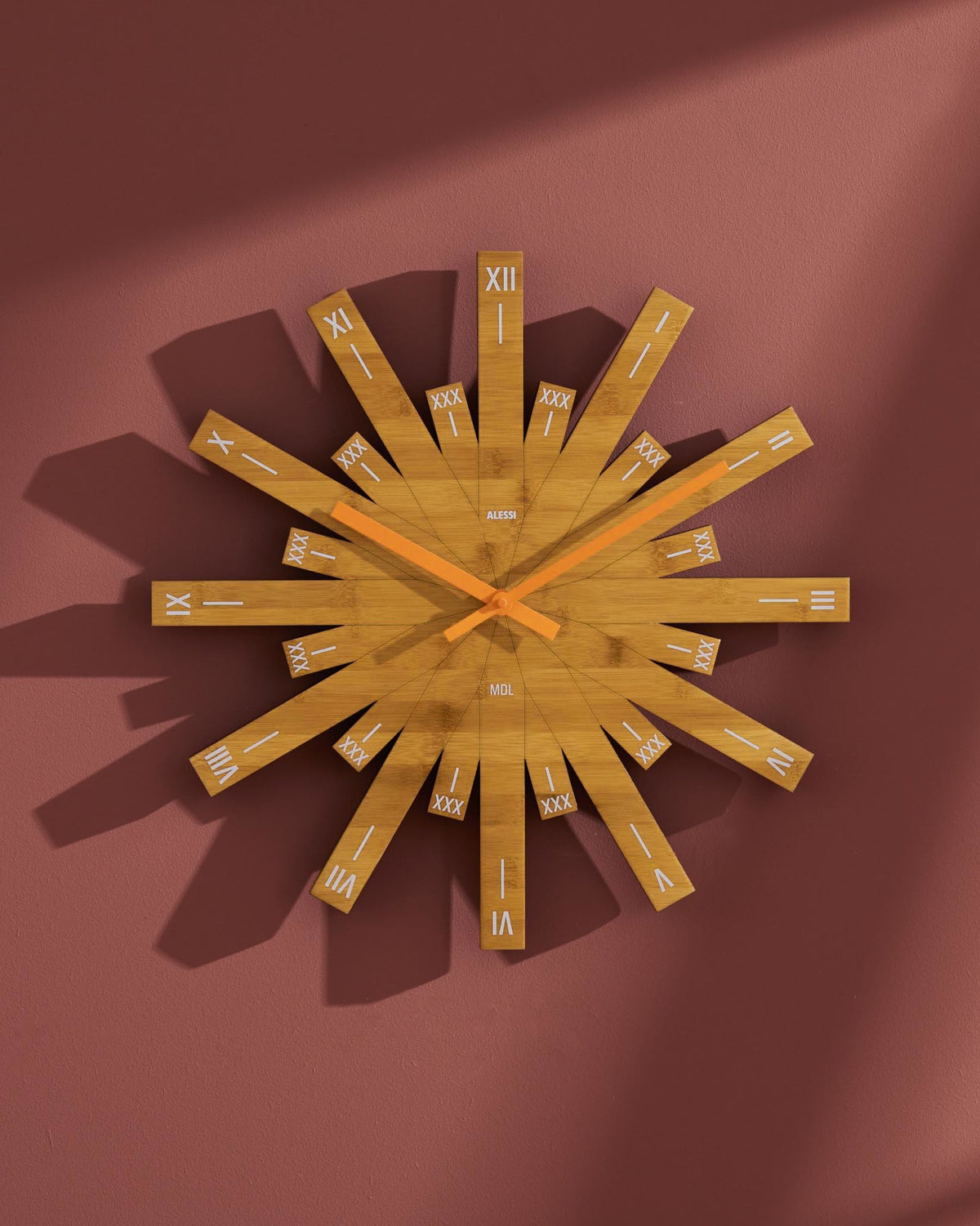 Light brown wooden starburst wall clock by Alessi, with orange hands and Roman numerals, on a reddish wall.