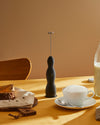 Black Pulcina handheld milk frother by Alessi with a ribbed design, standing on a yellow table beside a cup of frothed coffee and cake.