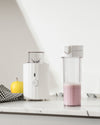 White ribbed blender by Alessi with clear top blending cup, alongside a clear bottle containing pink smoothie.