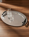 Silver oval serving tray by Alessi with a studded edge and black ergonomic handles, holding a glass water bottle and drinking glass.