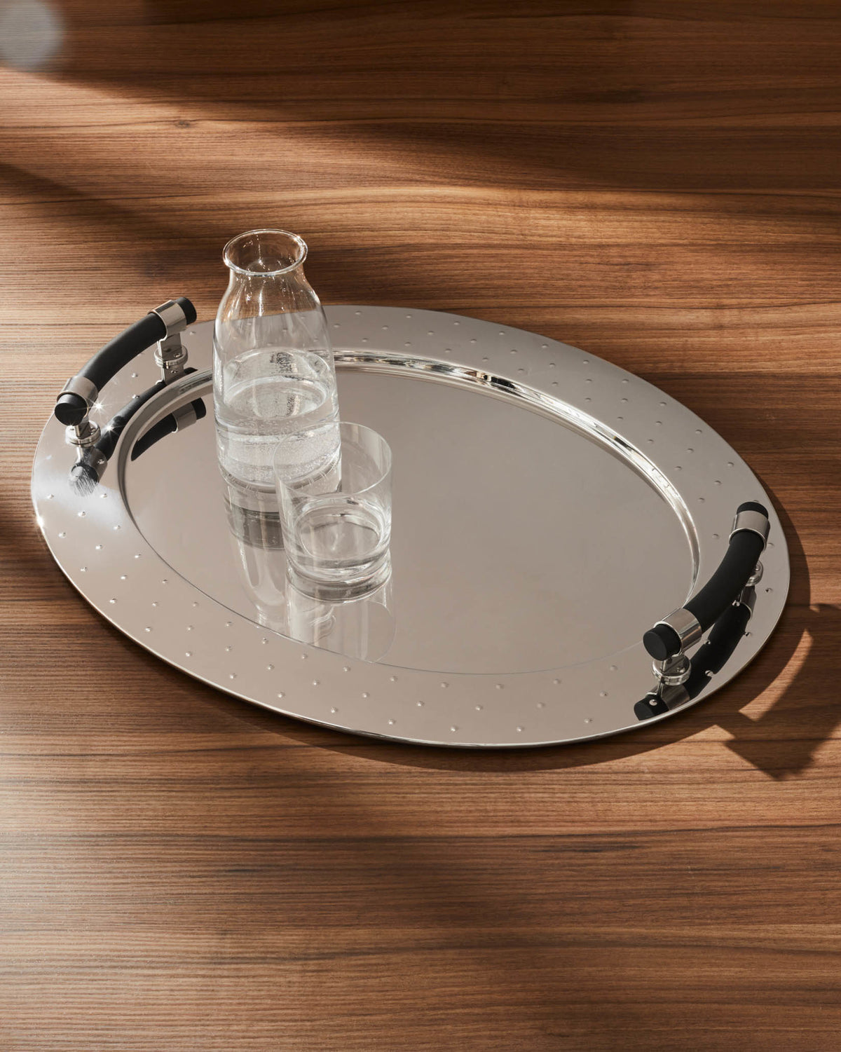 Silver oval serving tray by Alessi with a studded edge and black ergonomic handles, holding a glass water bottle and drinking glass.