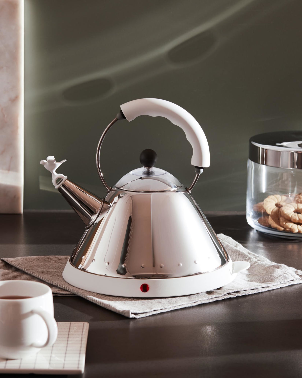 Silver conical kettle with white handle and base by Alessi, featuring a bird-shaped whistle on the spout and a red indicator light.