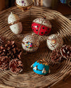 Blue spherical ornament with white bees and gold accents by Alessi, resting in a woven basket with pinecones.