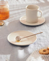 Acacia honey dipper, metallic with a spherical design, by Alessi, resting on a white plate with honey, a glass jar, white cup, and cookies.