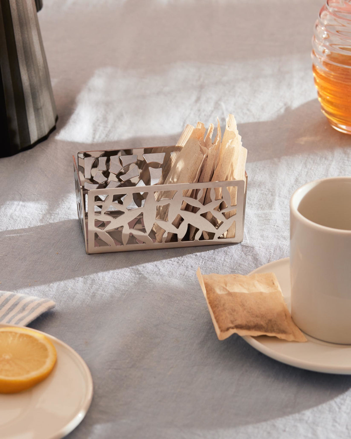 Silver metal tea bag holder by Alessi, featuring a modern cutout cactus design, filled with tea bags on a blue linen tablecloth.
