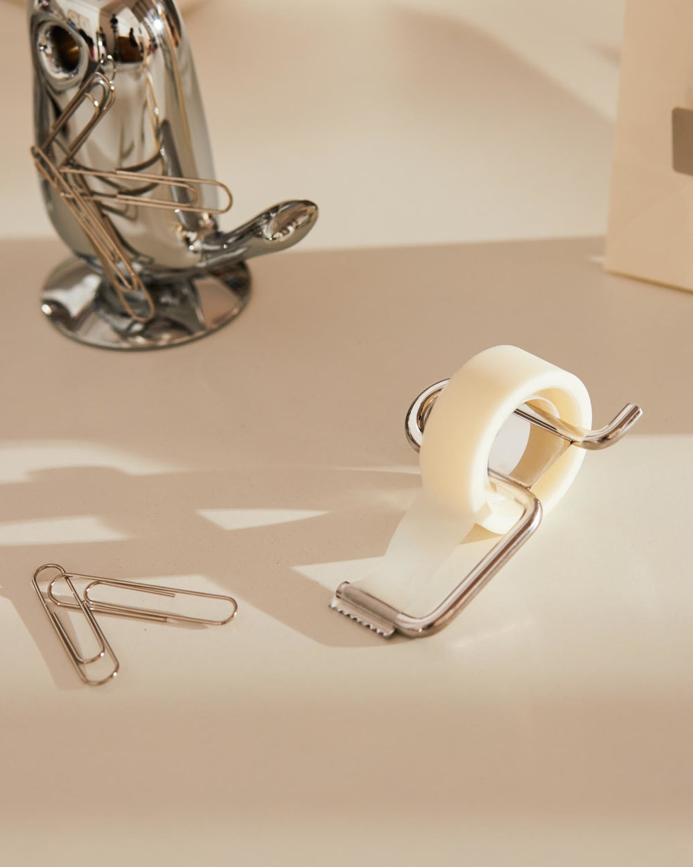 Filo tape dispenser with a chrome wire frame and cream tape by Alessi, angled on a desk next to paper clips and a chrome fish holder.