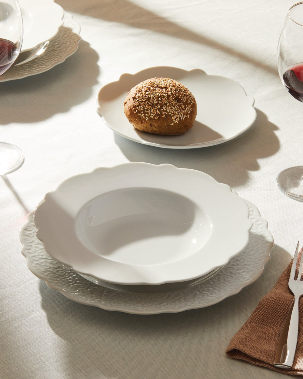 White dinnerware set by Alessi, featuring scalloped edges, an embossed underplate, and a soup bowl, with a bread roll on a sunlit table.