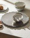 Silver scalloped serving platter by Alessi, with embossed scroll pattern, displayed with white bowls and ornate spoons.