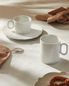 White porcelain cup and saucer set by Alessi, with an ornate embossed pattern on the saucer, on a white tablecloth.