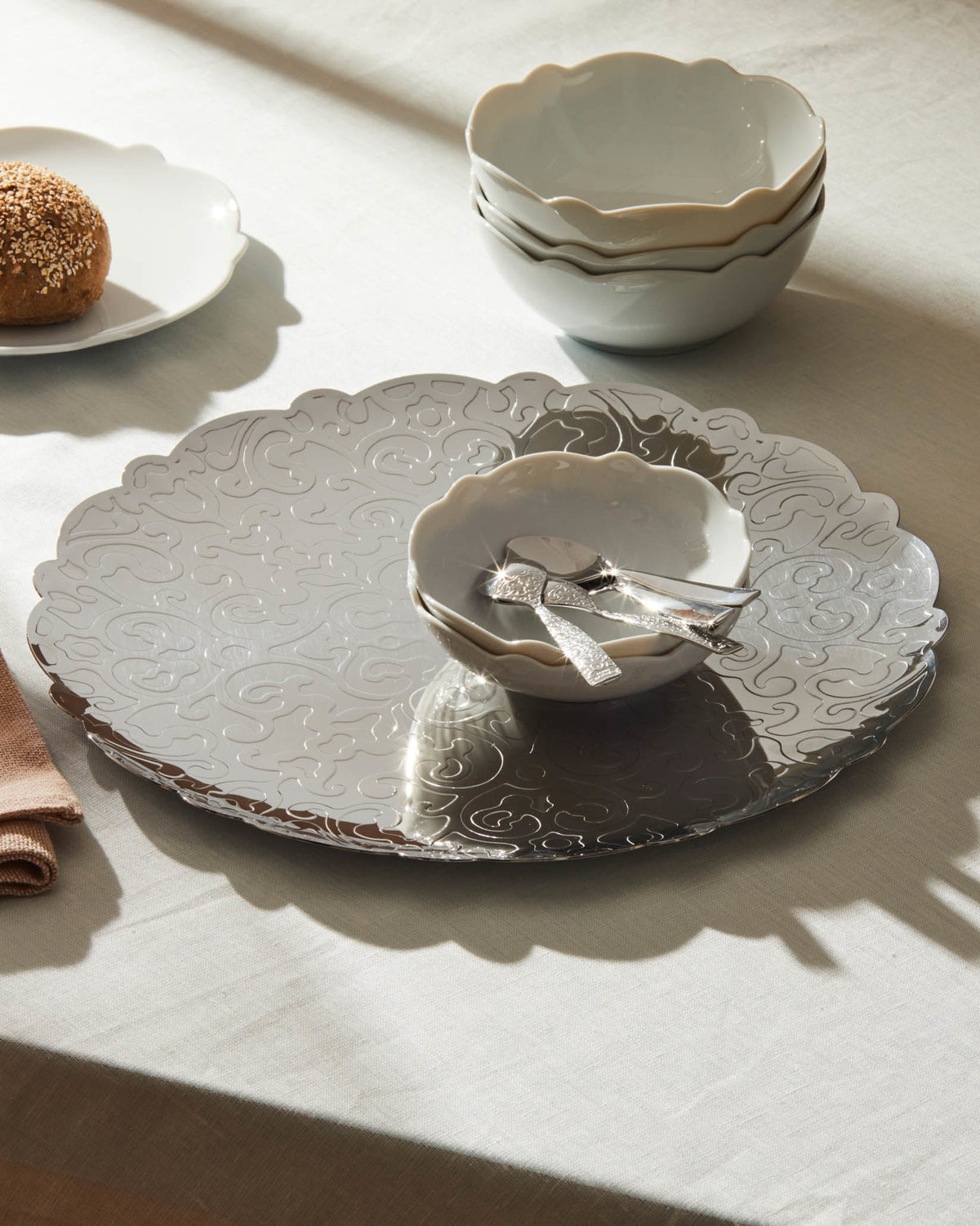 Silver platter by Alessi, with a scalloped edge and ornate embossed swirling pattern, displaying white bowls and a bread roll.