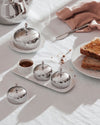 Polished silver lidded serving dishes by Alessi on white embossed ceramic trays with jam and toast.