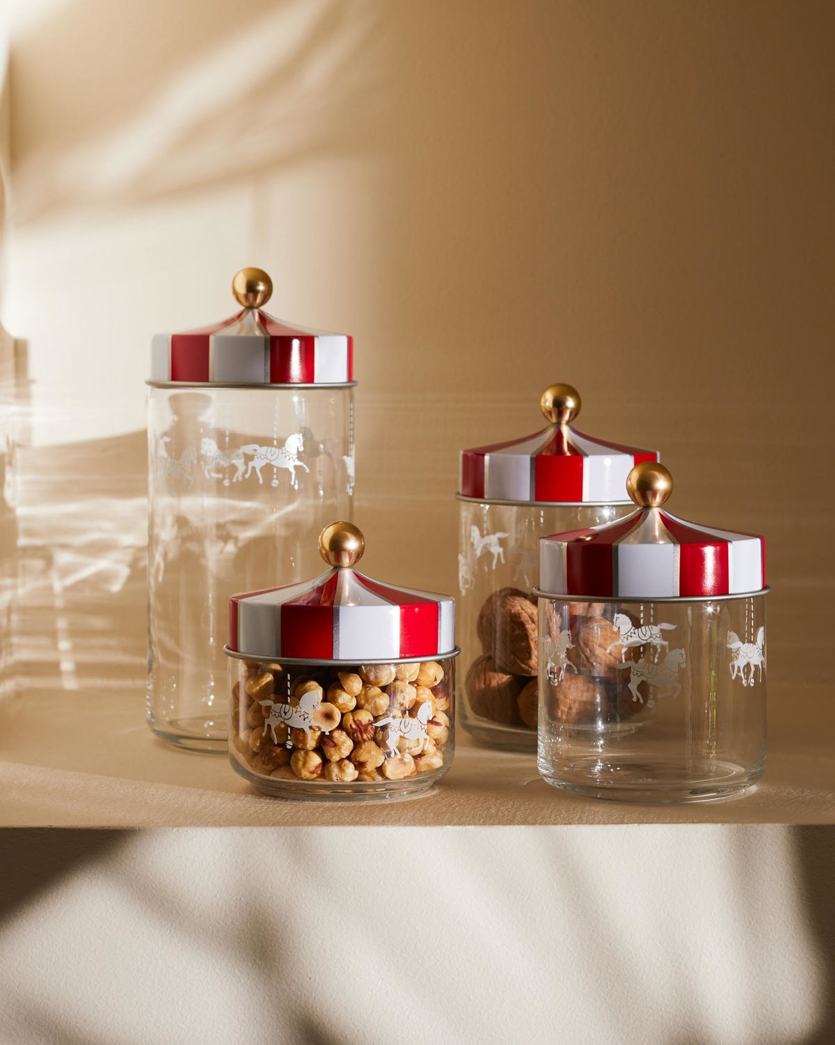 Clear glass storage jars by Alessi with red and white striped circus tent lids, golden handles, and carousel horse etching.