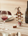 Jester-shaped corkscrew by Alessi, silver and multi-colored with a harlequin pattern, on a table next to a wine glass.
